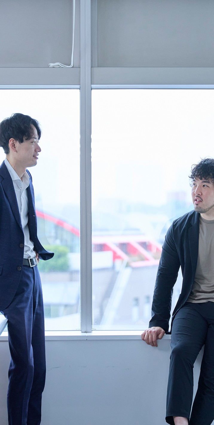 窓際に立つ2人のビジネスマン。会議室のような場所で、打ち合わせをしている様子。ホワイトボードも見える。