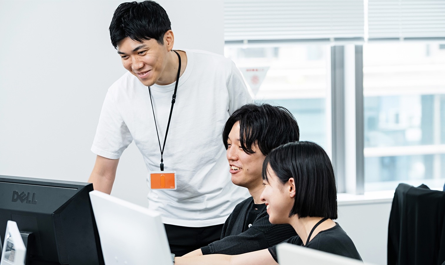 オフィスで、笑顔の男性が2人の同僚と共にパソコンに向かって作業している様子。チームワークや共同作業の様子を表す画像。