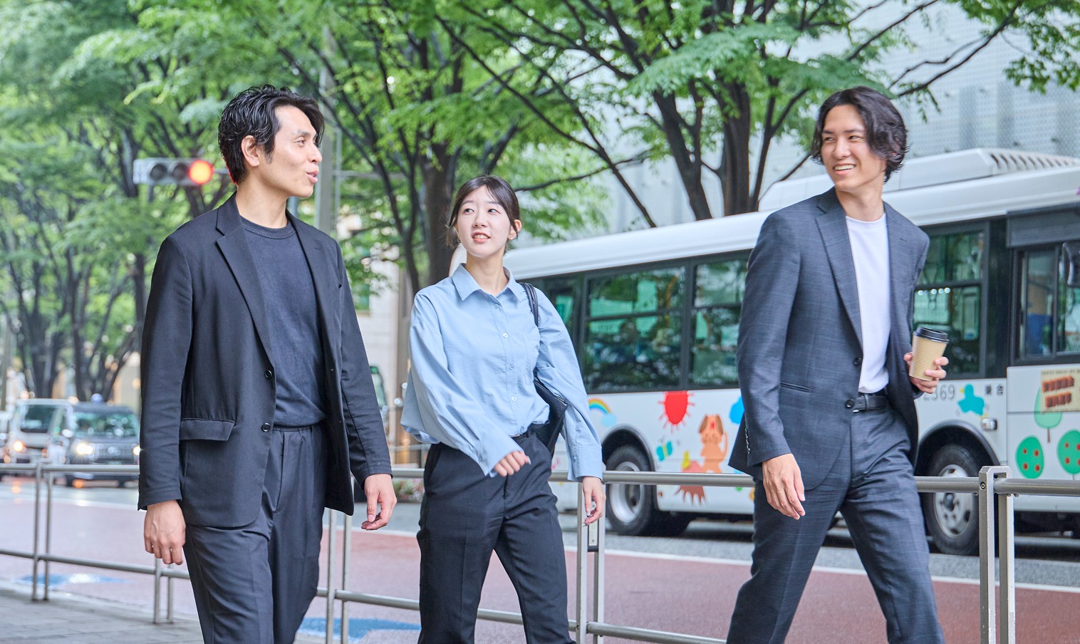 都会の街路で、ビジネスカジュアルな服装の男女3人が談笑しながら歩いている様子。背景にバスと緑の木々が見える。